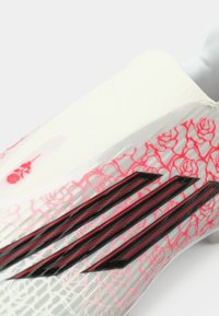 Ποδοσφαιρικό παπούτσι με λευκή βάση, ροζ λουλουδάτο μοτίβο και μαύρες λεπτομέρειες, συμπεριλαμβανομένων τριών καμπυλωτών ραβδώσεων στο πλάι.