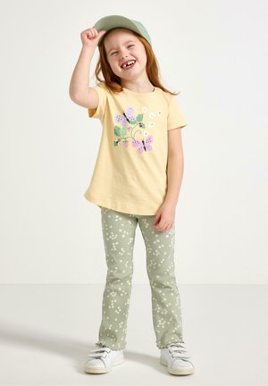 Smilende pige iført en gul skjorte med sommerfugleprint, grønne bukser med blomsterprint, hvide sneakers og en lysegrøn kasket, stående på en ensfarvet baggrund.