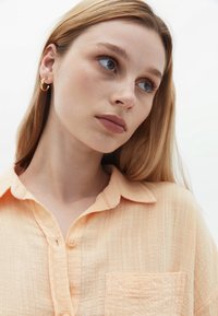 Camicia leggera color pesco, con bottoni, caratterizzata da una trama testurizzata e una tasca applicata. Il modello indossa orecchini a cerchio d'oro piccoli.
