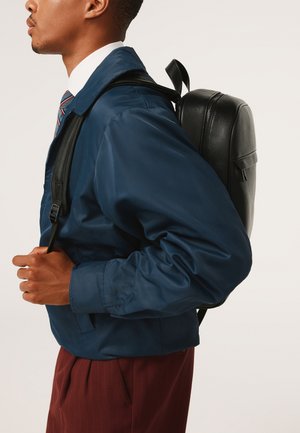 Mies tummansininen takki ja raidallinen solmio, kantaa mustaa nahkareppua selässään, rajattu hartioista lantiolle, neutraali tausta.