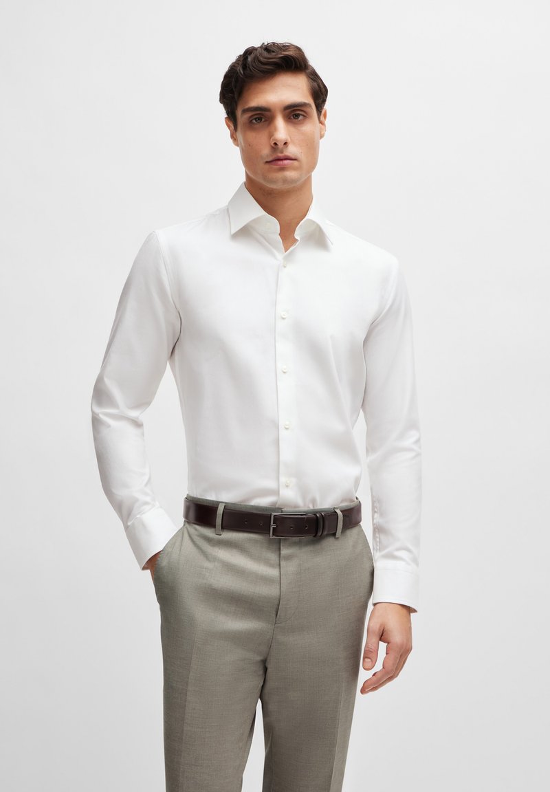 Witte langslavende shirt gemaakt van een gladde stof, met een klassieke kraag en knoopsluiting, gecombineerd met lichtgrijze broek en een bruine riem.