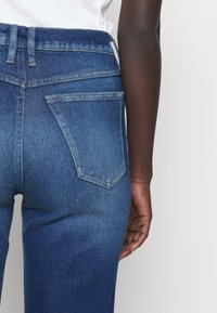 Jeans de mezclilla en azul oscuro, con una cintura alta, bolsillos traseros con solapas puntiagudas y detalles de costura visibles. Textura suave en toda su superficie.