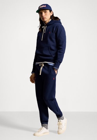 Hoodie bleu marine avec cordons blancs et poche kangourou, associé à un pantalon de survêtement assorti portant un logo. Baskets blanches et casquette.
