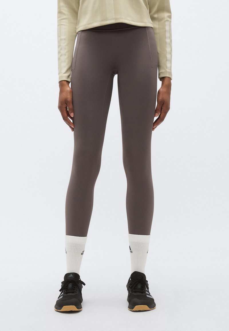 Leggings marron moulants avec une texture lisse, associés à des chaussures de sport noires et des chaussettes blanches montantes, avec des détails de couture subtils.