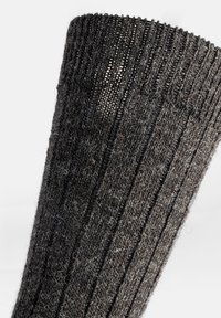 Calcetines de lana gris de punto con una textura acanalada, mostrando un ligero brillo y un patrón intrincado, con un puño reforzado y fibras sueltas.