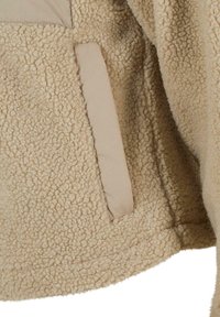 Bež sekcija sherpa flis jakne koja pokazuje teksturirano tkanje i okomiti, glatki otvor džepa sa strane.