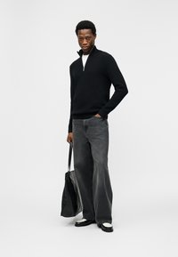 Homme portant un pull noir à demi-fermeture éclair, jean gris à jambes larges, chaussures noir et blanc, tenant un grand sac cabas noir, debout sur fond blanc.