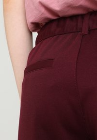 Burgundské kalhoty s texturovanou látkou a elastickým pasem, které mají malou boční kapsu a jemné švy.