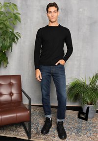 Suéter negro de manga larga, jeans azul ajustado, zapatillas negras. Silla de cuero marrón, planta verde, alfombra con estampado, fondo de pared gris.