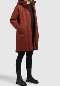 Brun vandtæt parka med hætte, frontlynlås og to sidelommer, parret med sorte leggings og ankelstøvler. Glat tekstur.
