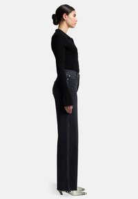 Top a maniche lunghe nere a costine abbinato a jeans scuri a vita alta. Modella indossa stivali alla caviglia a punta in pelle di serpente. Silhouette corporea verticale.