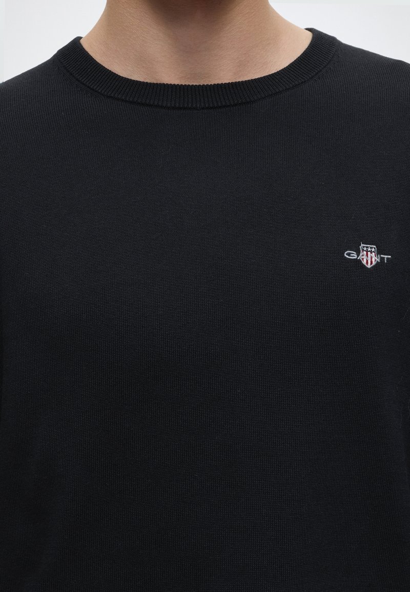 Black crew neck sweater with small GANT logo featuring shield and stars on the upper left chest, worn by a person shown from neck to mid-torso.