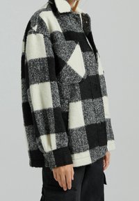 Svart och vit rutig jacka gjord av tjockt, texturerat tyg. Har krage och framfickor, med varierande fyrkantiga mönster.