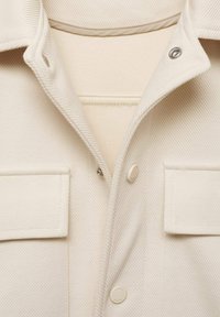 Light beige jacket with a textured fabric, featuring a pointed collar, metal snaps, and two front pockets. Accentuated by smooth stitching.