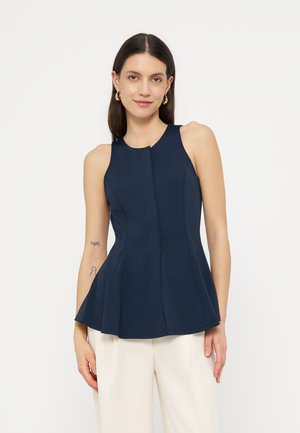 Femme portant un haut péplum sans manches bleu marine et un pantalon blanc, debout devant un fond clair uni, faisant face à l'avant avec une expression détendue.