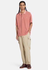 Lys pink polo shirt med knapkrave, parret med beige cargo bukser med sidelommer og brune sko med snørebånd.