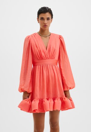 Femme portant une robe rose corail avec des manches longues bouffantes, un col en V profond, une taille cintrée et un ourlet à volants sur un fond clair uni.