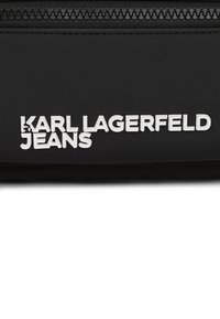 Svart stoffpose med teksturert overflate og en fremtredende hvit logo som leser "KARL LAGERFELD JEANS." Glidelås lukking på toppen.