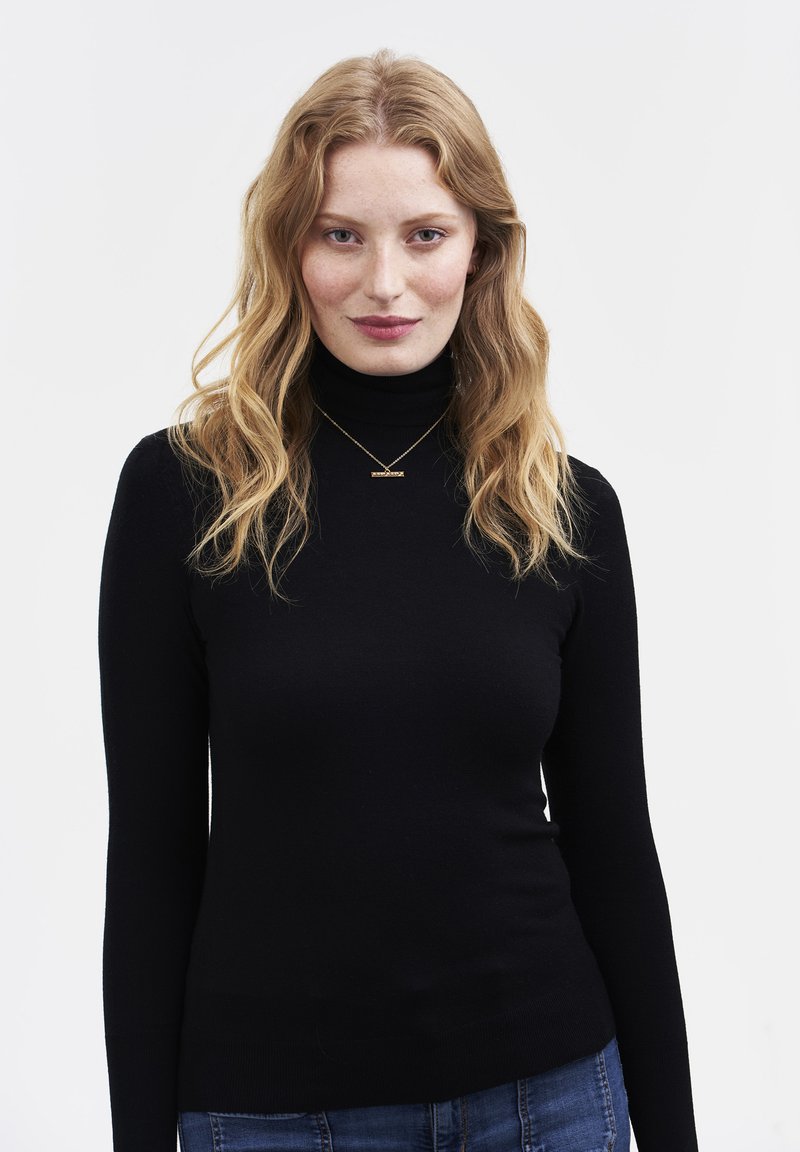 Sieviete ar viļņainiem blondiem matiem, tērpusies melnā rolkrekla džemperī, zilās džinsās un zelta kaklarotā, stāvot pret vienkāršu baltu fonu.