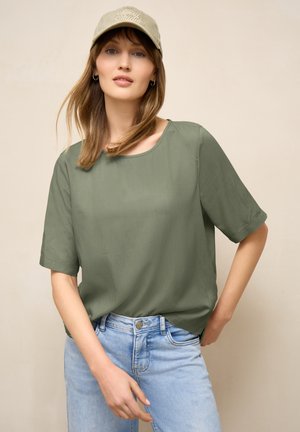 Kvinde med lyst brunt hår iført en olivengrøn top med korte ærmer, lyseblå jeans og en beige kasket mod en ensfarvet baggrund.