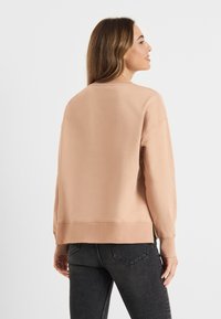 Jasno brzoskwiniowy sweatshirt z długimi rękawami, ściągaczami przy mankietach i na dole. Posiada luźny krój i boczne rozcięcia dla swobody ruchów, idealnie pasujący do ciemnych dżinsów.