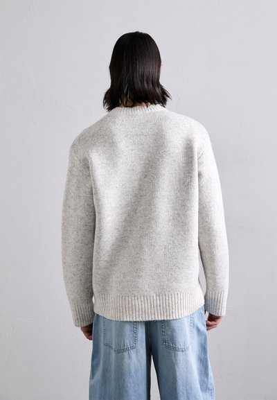 Világosszürke kötött pulóver texturált felülettel. Bordás mandzsetták és szegély. Feslett, laza világoskék farmernadrág felett viselve.