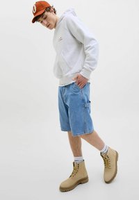 Grå hættebluse, blå denim shorts med lomme, beige snørebåndsstøvler og hvide sokker med tekst. Minimalistisk baggrund.