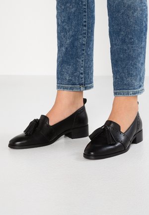 Mocassins à glands en cuir noir portés avec un jean bleu court sur un sol clair, fond blanc