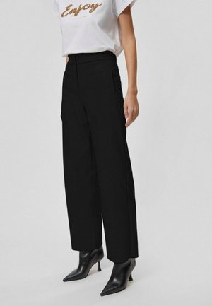 Žena nosí černé široké kalhoty s vysokým pasem, bílou krátkým rukávem tričko s nápisem „Enjoy“ a černé boty na podpatku s špičatou špičkou.