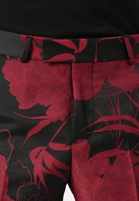 Schwarze Hose mit einem strukturierten roten Blumenmuster, flachem Bund und Gürtelschlaufen. Der Stoff hat einen leichten Glanz und aufwendige Details.