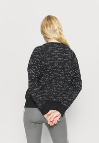 Sudadera negra con un patrón de triángulos blancos, con cuello redondo y puños acanalados. Se lleva con leggings grises, vista desde atrás.