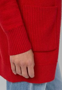 Ręka spoczywająca na biodrze, ubrana w czerwoną dzianinową bluzę z prążkowanymi mankietami i kieszenią, w połączeniu z jasnoniebieskimi dżinsami.