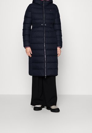 Manteau rembourré bleu marine avec capuche, taille ceinturée et fermeture éclair. Il présente des coutures horizontales et une doublure intérieure contrastante. Associé à un pantalon noir.