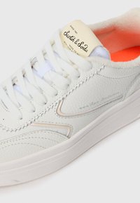 Weiße Ledersneaker mit weißen Schnürsenkeln, sichtbarem Markenlabel "Scotch & Soda" auf der Zunge und orangefarbigem Innenfutter.