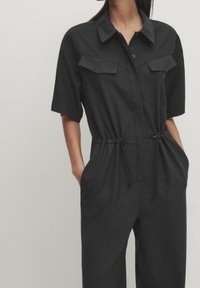 Black short-sleeve jumpsuit with a button-up collar, two chest pockets, adjustable drawstring waist, and a relaxed fit. Soft fabric texture.