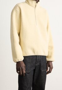 Pullover di pile giallo con colletto alto e mezza zip; presenta spalle cadenti e polsini a costine, indossato su jeans denim scuri.