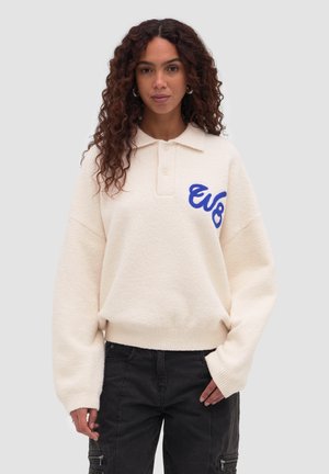 Femme aux cheveux longs et bouclés portant un pull crème avec des lettres bleues sur la poitrine et un pantalon noir, sur un fond clair uni.