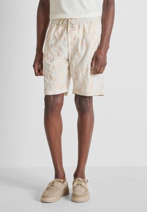 Uomo che indossa pantaloncini beige con fantasia floreale, top in maglia color crema e scarpe beige con lacci su sfondo semplice.
