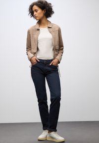 Světle hnědá semišová bunda, bílá košile, tmavě modré džíny a bílé tenisky. Bunda má stojáčkový límec a dvě kapsy.