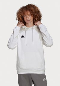 Hvit hettegenser laget av bomull, med kengurulomme og justerbar snor. Svart Adidas-logo på venstre bryst.