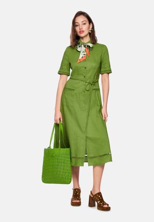 Woman wearing a green buttoned midi dress with tied waist, floral neck scarf, carrying a green woven tote, and brown heeled sandals.