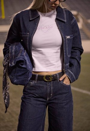 Femme portant une veste en jean foncée et un jean, top court blanc avec motif en strass, tenant une veste en jean pliée avec une écharpe noire à motif.