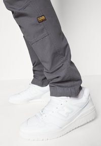 Šedé cargo kalhoty z texturované látky mají boční kapsu a malé logo G-Star. Doplňují je bílé sportovní tenisky s perforovanými detaily.