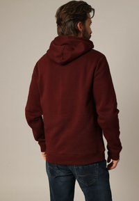 Bordeaux hoodie met een zachte textuur, kangoeroezak en geribbelde manchetten. Draag met blauwe jeans, wat een casual uitstraling van achteren laat zien.