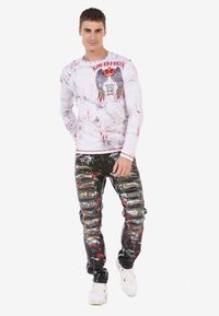 Langärmliges weißes Hemd mit roten und schwarzen Farbspritzern, das grafische Flügel und Text zeigt. Schwarze abgetragene Jeans mit bunten Farbschattierungen.