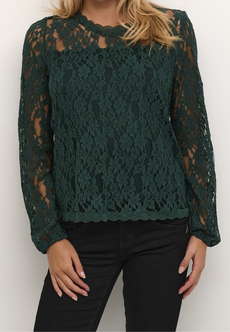 Femme portant un haut à manches longues en dentelle florale vert foncé sur une couche de base unie foncée, associé à un pantalon noir.