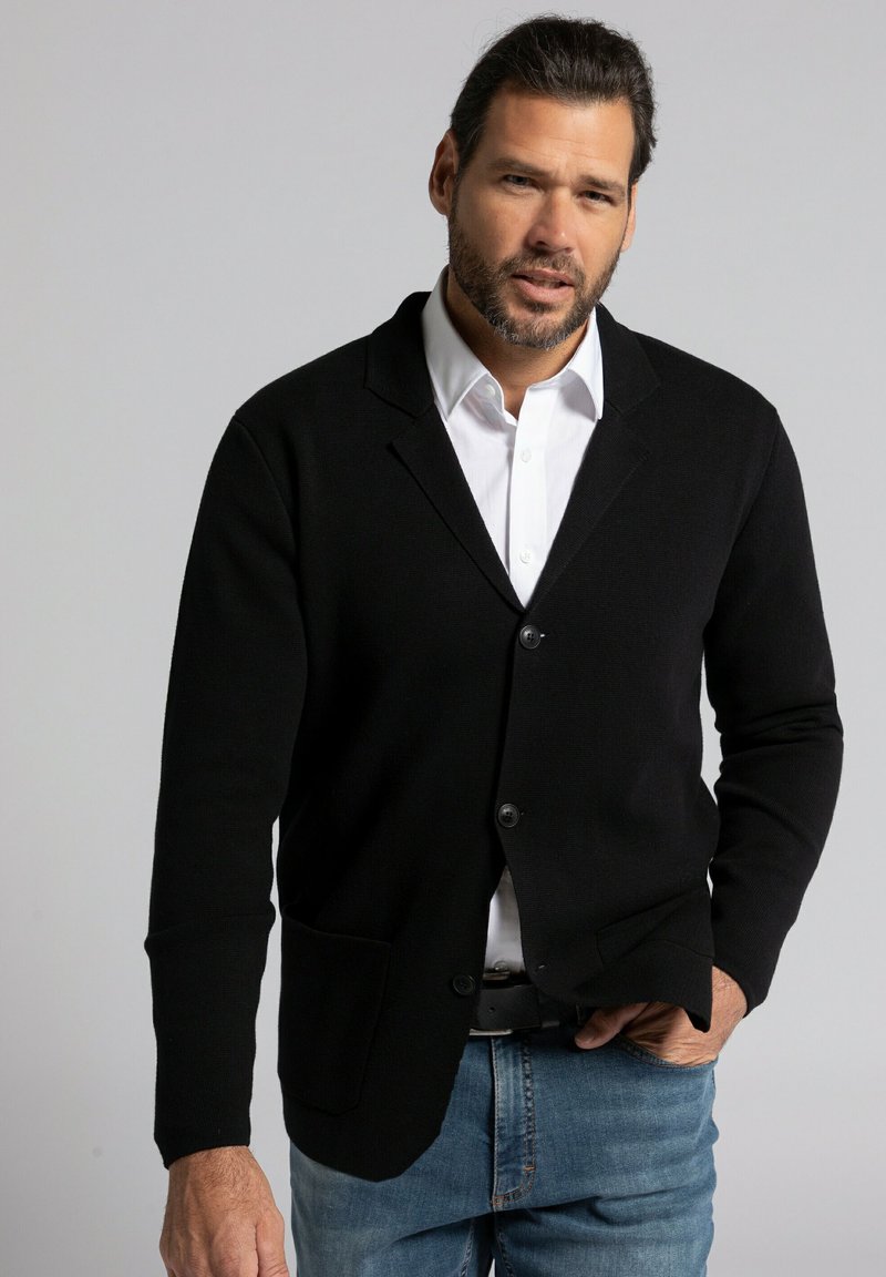 Schwarze Strickjacke mit schlankem Schnitt, mit einem Reverskragen, zwei vorderen Taschen und Knopfverschluss, kombiniert mit einem weißen Hemd und Jeans.
