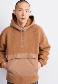 Pulôver de fleece castanho com gola com fecho e bolso tipo mochila, fabricado em tecido texturizado. Apresenta um detalhe liso em tom bege na área do bolso.