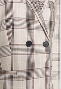 Blazer doppiopetto beige con pattern scozzese marrone e giallo. Presenta bottoni neri e un tessuto testurizzato con una vestibilità su misura.