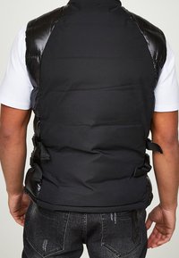 Veste matelassée noire avec des panneaux d'épaule brillants, une texture douce et des sangles ajustables sur les côtés. Portée par-dessus un t-shirt blanc, avec un dos uni.